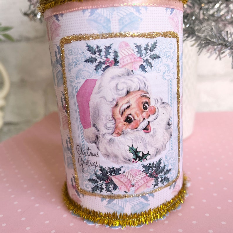 K&ouml;nnte beinhalten: Eine rosafarbene und blaue Weihnachtsdose mit goldener Glitzerverzierung. Die Dose zeigt eine Vintage-Illustration des Weihnachtsmanns mit dem Text "Christmas Greetings".