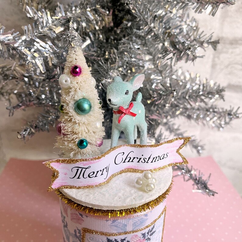 K&ouml;nnte beinhalten: Eine wei&szlig;e Flasche mit einem rosa und goldenen Banner, auf dem "Frohe Weihnachten" steht. Die Flasche ist mit einem wei&szlig;en Flaschenb&uuml;rstenbaum mit rosa und blauen Ornamenten, einer hellblauen Hirschfigur mit einer roten Schleife und wei&szlig;en Perlen verziert.