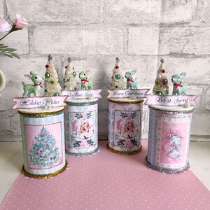 K&ouml;nnte beinhalten: Vier dekorierte Blechdosen mit pinkfarbenen und blauen Weihnachtsmotiven. Jede Dose hat ein anderes weihnachtliches Bild und einen anderen Text, darunter "Holiday Wishes", "Christmas Wishes", "Merry Christmas" und "Let it Snow".