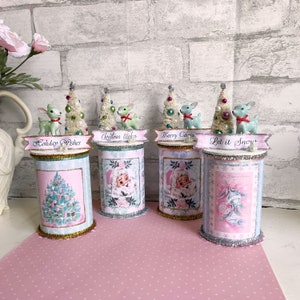 K&ouml;nnte beinhalten: Vier dekorative Weihnachtsbeh&auml;lter in einem rosa-blauen Farbschema. Jeder Beh&auml;lter zeigt eine andere Weihnachtsszene und eine kleine Keramik-Rehfigur auf der Oberseite. Die Beh&auml;lter sind mit "Holiday Wishes", "Christmas Wishes", "Merry Christmas" und "Let it Snow" beschriftet.