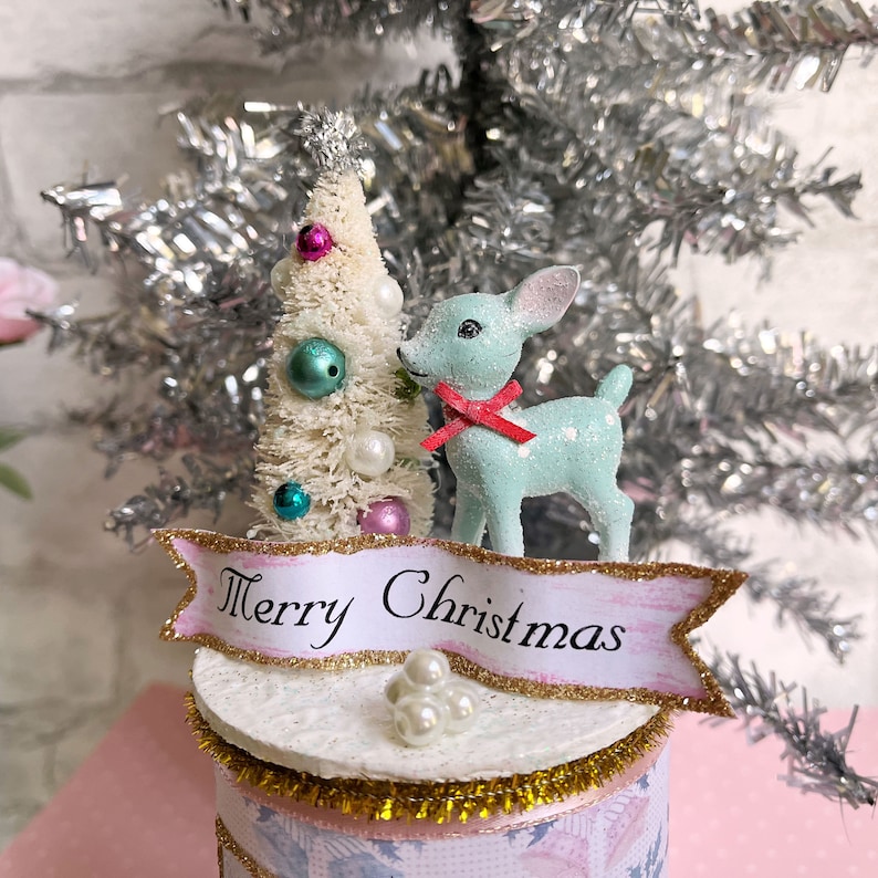 K&ouml;nnte beinhalten: Ein kleiner wei&szlig;er Weihnachtsbaum mit rosa und blauen Ornamenten, eine hellblaue Hirschfigur mit einer roten Schleife und ein Banner mit der Aufschrift "Frohe Weihnachten" auf einem wei&szlig;en Sockel mit goldenem Glitzer.