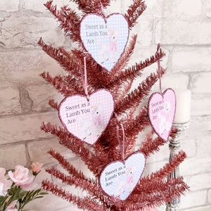 Puede incluir: Tres adornos en forma de corazón rosa y azul con purpurina y un diseño de cordero blanco. Cada adorno tiene el texto "Sweet as a Lamb You Are" impreso en él.