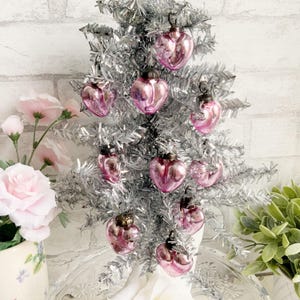 以下が含まれることがあります： ピンクのガラス製のハート型のオーナメントで飾られた、小さな銀色のクリスマスツリー