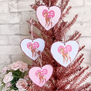 Puede incluir: Cuatro adornos en forma de corazón rosa y azul con un patrón de vichy blanco y una ilustración de un ciervo. Cada adorno tiene el texto "my deer" impreso en él.