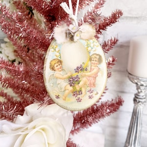May include: A vintage Easter egg ornament with a white background and two angels holding purple flowers. The ornament is decorated with a white bow and has a white string for hanging.
