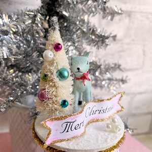 K&ouml;nnte beinhalten: Eine wei&szlig;e Flasche mit einem wei&szlig;en Flaschenb&uuml;rsten-Weihnachtsbaum, einer hellblauen Hirschfigur und einem rosa-goldenen Banner mit der Aufschrift "Frohe Weihnachten".