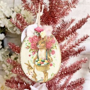 Op de afbeelding: Een vintage-stijl paaseieren ornament met een konijn dat een mandje met kleurrijke eieren vasthoudt. De ornament is versierd met roze rozen en heeft een witte achtergrond.