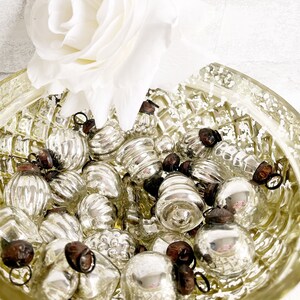 May include: A bowl filled with silver glass ornaments, some with a textured surface, others with a smooth surface. The ornaments are in a variety of shapes and sizes. The bowl is sitting on a white surface.