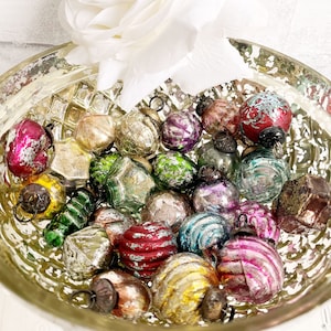 Puede incluir: Una colección de adornos navideños vintage en un cuenco de cristal decorativo. Los adornos son de varias formas y colores, incluyendo rosa, verde, dorado y rojo. Algunos tienen un acabado metálico, y otros son rayados o texturizados. Una rosa blanca está en el fondo.