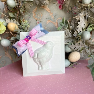 以下が含まれることがあります： 白い木製のブロックに、白いセラミック製の鳥のレリーフが施されています。ブロックは、ピンクと白のギンガムチェックのリボンと、青と白の花柄のリボンで飾られています。