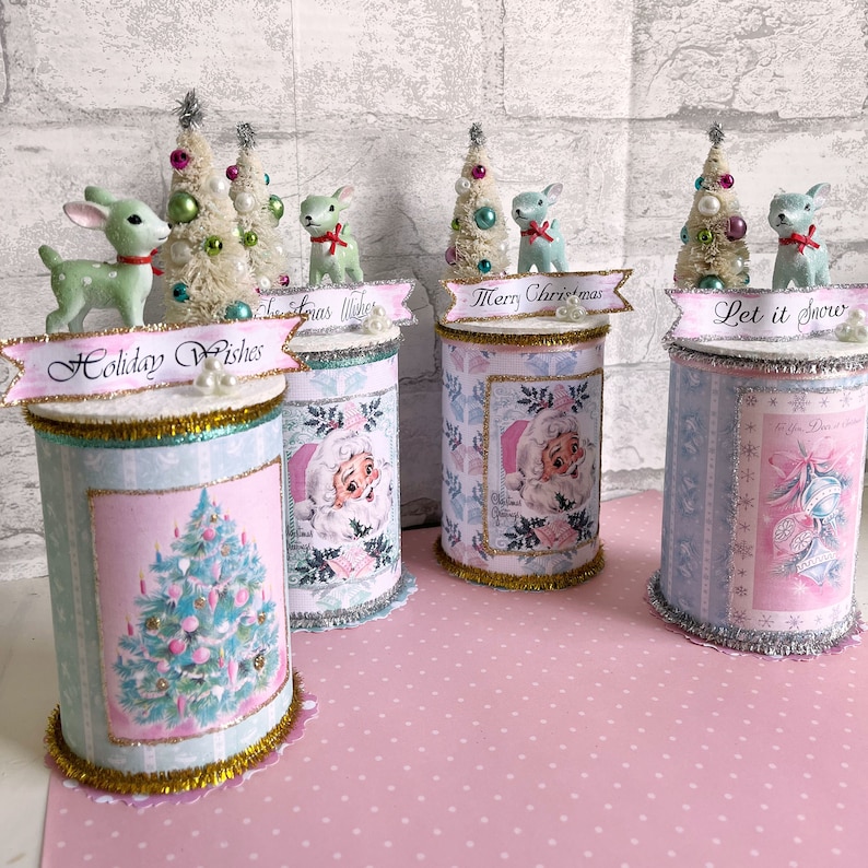 K&ouml;nnte beinhalten: Vier dekorierte Blechdosen mit Weihnachtsmotiven. Jede Dose hat ein anderes Design mit dem Weihnachtsmann, einem Weihnachtsbaum und einem Rentier. Die Dosen sind mit einem kleinen k&uuml;nstlichen Weihnachtsbaum, einer Rentierfigur und einem Banner mit einem Weihnachtsgru&szlig; verziert. Die Banner lauten "Holiday Wishes", "Merry Christmas", "Let it Snow" und "Christmas Wishes".