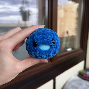 Op de afbeelding: Een blauw gehaakt speelgoed, bolvormig, met zwarte ogen en een lichtblauwe mond. Het speelgoed wordt in een hand gehouden, met een raam en een buitenscène op de achtergrond. Het speelgoed heeft een zacht, getextureerd uiterlijk.