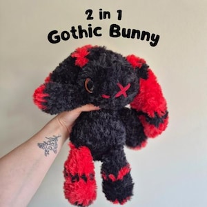 Könnte beinhalten: Ein schwarzes und rotes Gothic-Hasen-Plüschtier mit dem Text "2 in 1 Gothic Bunny". Der Hase hat rote Akzente an Ohren und Füßen sowie ein rotes Kreuz auf dem Gesicht. Der Hase wird von einer Person gehalten.