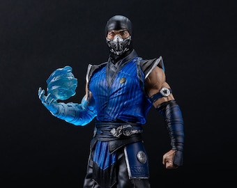 Könnte beinhalten: Eine blaue und schwarze Actionfigur von Sub-Zero aus der Mortal Kombat Videospielserie. Die Figur hält einen blauen Eisball in der rechten Hand. Die Figur steht auf einem schneebedeckten Sockel mit einem Mortal Kombat Logo.