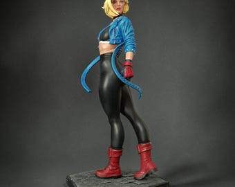 May include: A collectible figurine of a blonde character wearing a blue cropped jacket, black crop top, and black leggings. The figure features red gloves and red boots, standing on a gray hexagonal base with a textured stone-like surface.
