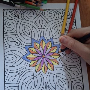May include: A person coloring a mandala design with colored pencils. The mandala features a central flower with blue, yellow, orange, and purple petals. The design is surrounded by intricate patterns.