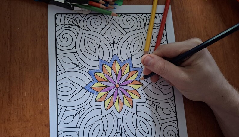 May include: A person coloring a mandala design with colored pencils. The mandala features a central flower with blue, yellow, orange, and purple petals. The design is surrounded by intricate patterns.