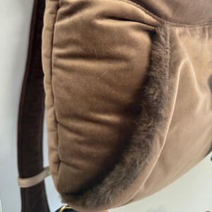 May include: A brown backpack with a soft, velvety texture. The edges are trimmed with a darker, furry material. The straps are a darker brown color, and the bag appears to be made of a plush material.