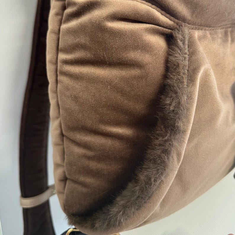 May include: A brown backpack with a soft, velvety texture. The edges are trimmed with a darker, furry material. The straps are a darker brown color, and the bag appears to be made of a plush material.
