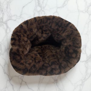 May include: A plush, brown pet bed with a leopard print pattern. The bed is circular and appears soft and comfortable, perfect for a small dog or cat. The faux fur material is a mix of dark and light brown tones.