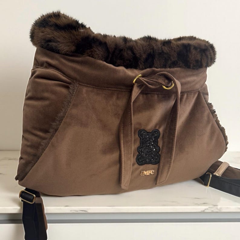 May include: Brown velvet pet carrier with a faux fur trim. Features a black sequined teddy bear applique and gold-toned hardware. Includes a black shoulder strap and the letters "MFC".