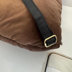 May include: A brown, plush pillow with a black leather strap and a gold-colored metal buckle. The pillow has a soft, velvety texture. The strap is attached to the pillow with a gold-colored metal buckle.