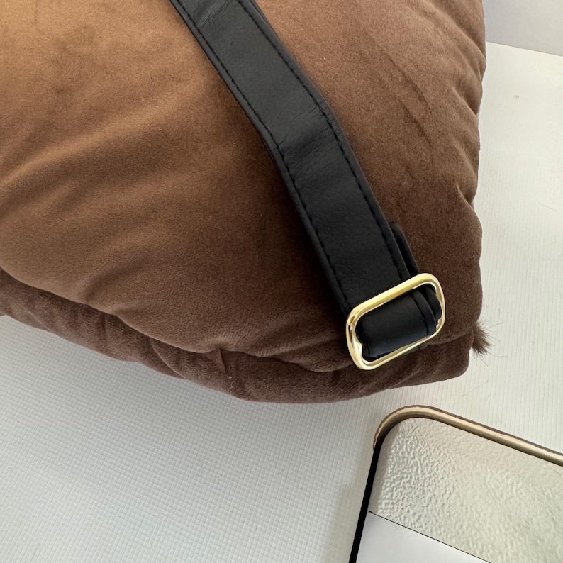 May include: A brown, plush pillow with a black leather strap and a gold-colored metal buckle. The pillow has a soft, velvety texture. The strap is attached to the pillow with a gold-colored metal buckle.