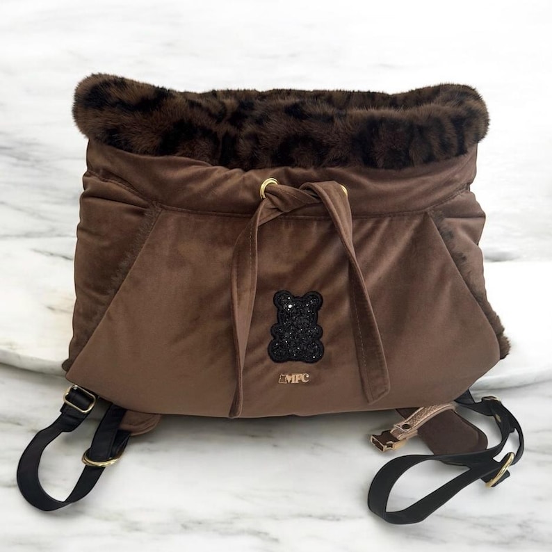 May include: A brown velvet pet carrier with a faux fur trim. The carrier features a drawstring closure, gold-tone hardware, and a black sequined bear emblem. Adjustable black straps and a small gold-tone strap are also visible.