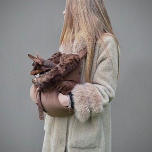 May include: A light brown pet carrier with a brown and faux fur interior, held by a person wearing a long, light beige coat with faux fur cuffs. A small brown dog is visible in the carrier.