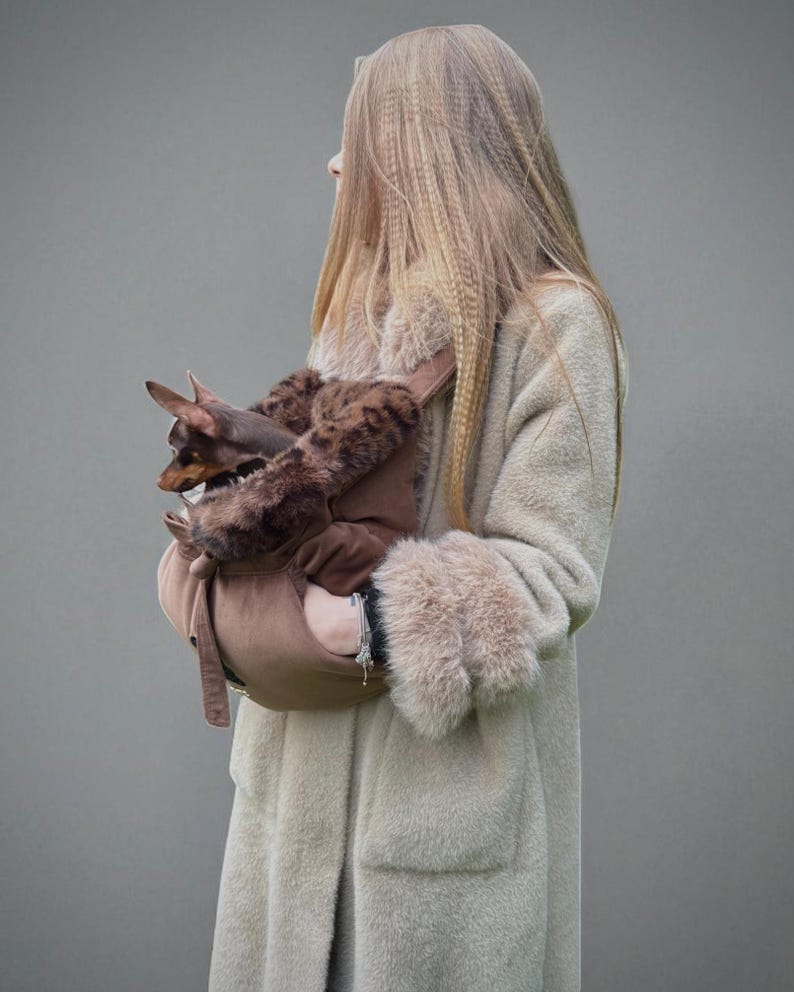 May include: A light brown pet carrier with a brown and faux fur interior, held by a person wearing a long, light beige coat with faux fur cuffs. A small brown dog is visible in the carrier.