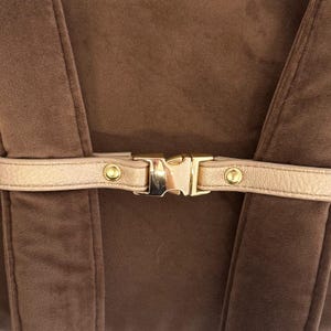 May include: Close-up of a brown velvet fabric with a gold-colored buckle and strap. The strap is a light gold color and is secured with a gold-colored metal buckle. The velvet fabric is a rich brown color.