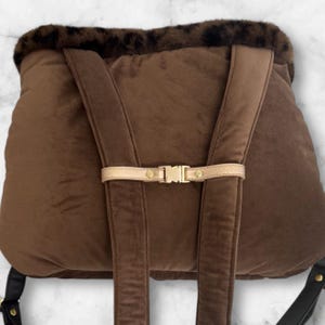 May include: A brown backpack with adjustable shoulder straps and a gold-colored buckle closure. The top of the backpack has a faux fur trim with a leopard print design. The straps are brown and the hardware is gold.