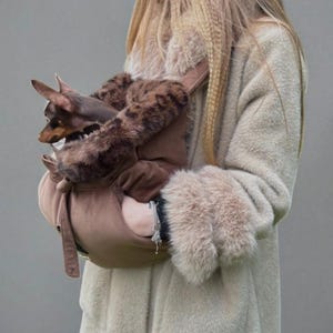 Könnte beinhalten: Eine braune Haustiertasche mit Kunstfellbesatz, in der sich ein kleiner braun-schwarzer Hund befindet. Die Tasche hat einen Schultergurt. Die Person trägt einen hellbraunen Mantel mit Pelzbündchen. Die Tasche ist für kleine Haustiere konzipiert.