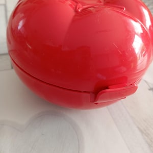 Peut inclure: Un récipient rouge vif en forme de tomate avec une finition brillante. Le récipient a un couvercle à charnière et une fermeture à fermoir. Le design imite la forme d'une tomate, avec des sections segmentées et un détail en forme de tige sur le dessus.