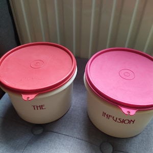 Peut inclure: Deux récipients de conservation des aliments blanc cassé avec des couvercles roses. L'un porte l'inscription "THE", l'autre "INFUSION". Les récipients sont ronds et semblent être en plastique.