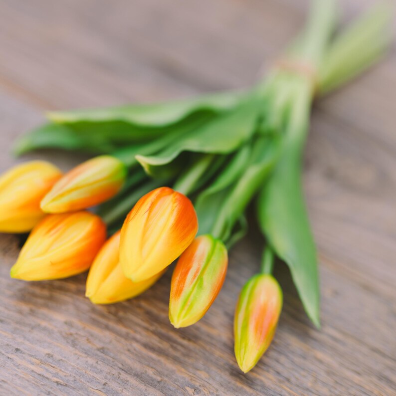 May include: A bouquet of tulips with yellow and orange petals and green leaves. The flowers are arranged on a wooden surface, creating a natural and vibrant display. The tulips are in full bloom, showcasing their colorful petals.