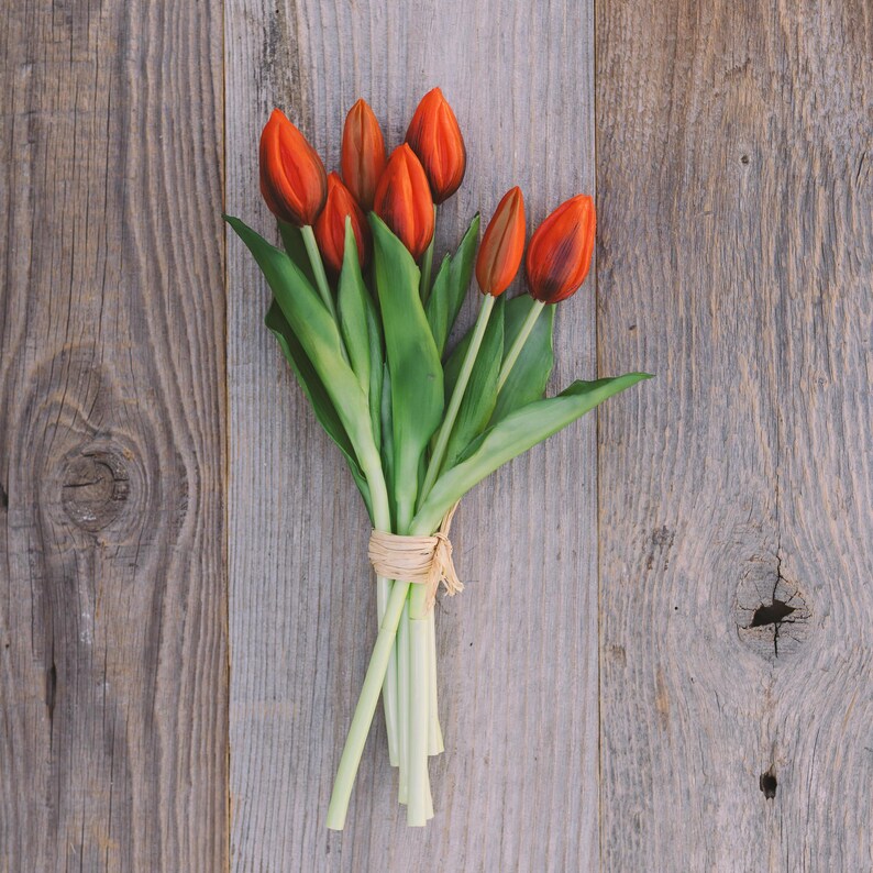 May include: A bouquet of orange tulips with dark red tips and green leaves, tied with a natural raffia ribbon. The flowers rest on a weathered, gray wooden surface, creating a rustic aesthetic.