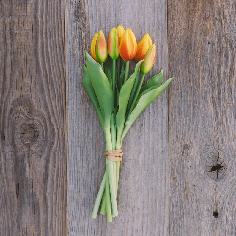 May include: A bouquet of yellow and orange tulips with green leaves, tied with twine, rests on a weathered wooden surface. The tulips are in full bloom, showcasing their vibrant colors. The wooden background has a rustic, aged appearance.