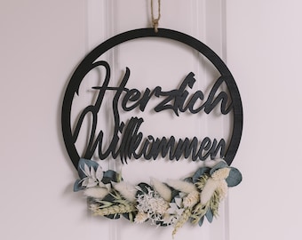 Op de afbeelding: Zwart houten welkomstbord met de woorden "Herzlich Willkommen" in sierlijke letters. Het bord heeft een cirkelvorm en is versierd met gedroogde bloemen en groen, en hangt aan een natuurlijke touw.