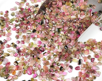 May include: A large pile of small, tumbled, pink, green, and black tourmaline gemstones. The gemstones are scattered on a white surface.