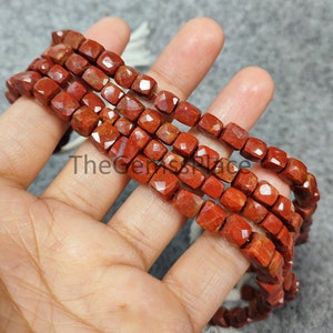 Puede incluir: Una hebra de cuentas de jaspe rojo, cada cuenta está facetada y tiene forma cuadrada. Las cuentas están enhebradas en un cordón blanco.