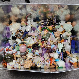 May include: A metal container overflowing with a variety of colorful gemstones, crystals, and beads. The collection includes amethyst points, quartz clusters, and beaded necklaces in shades of purple, pink, green, and gold. A diverse assortment of natural stones.