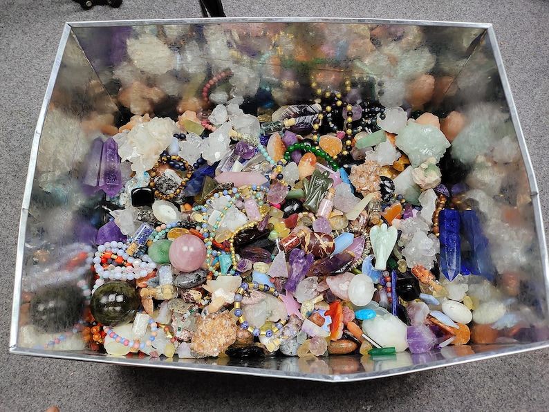 May include: A metal container overflowing with a variety of colorful gemstones, crystals, and beads. The collection includes amethyst points, quartz clusters, and beaded necklaces in shades of purple, pink, green, and gold. A diverse assortment of natural stones.