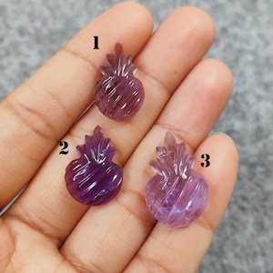 Pode incluir: Três pingentes de pedra preciosa de ametista esculpidos em forma de abacaxi. Cada pingente tem um tom diferente de roxo, com detalhes esculpidos intrincados. Os pingentes são segurados em uma mão contra um fundo neutro.