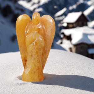 May include: A carved angel figurine made of yellow citrine crystal. The angel is standing on a white surface with a snowy background.