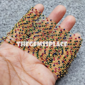 May include: A close-up of a gold chain with red, blue, and green beads. The chain is folded in half and held in a hand. The text "THEGEMSPLACE" is visible on the chain.