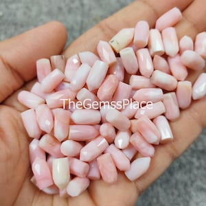 Könnte beinhalten: Nahaufnahme eines Stapels rosa und weißer Opalperlen. Die Perlen sind facettiert und haben eine glatte, polierte Oberfläche. Die Perlen sind alle ungefähr gleich groß und gleich geformt. TheGemsPlace ist auf dem Bild gedruckt.