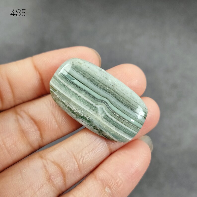 May include: A green and white banded agate cabochon with a smooth, polished surface. The stone has a natural, organic pattern.