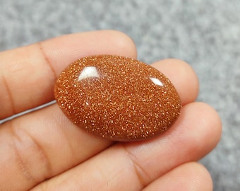 May include: A close-up of a smooth, oval-shaped, orange sunstone cabochon. The stone has a sparkly, glittery texture.