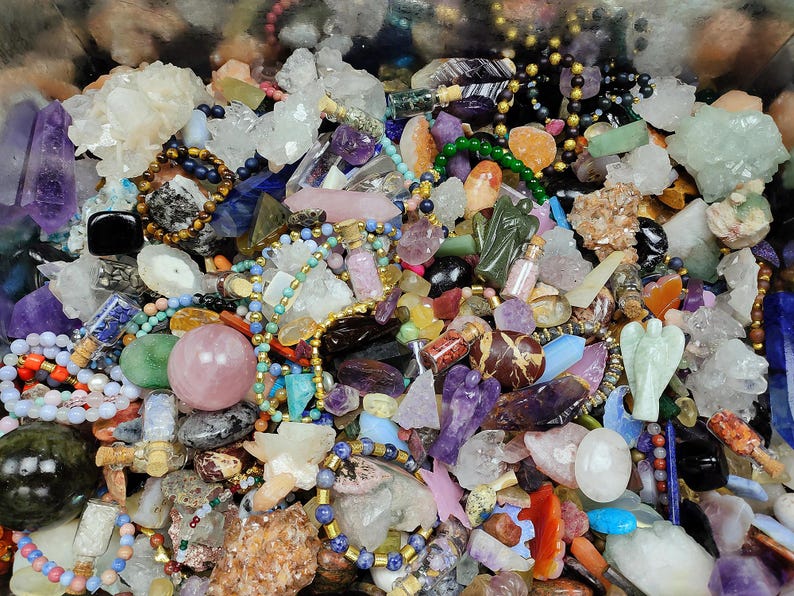 May include: A collection of various gemstones and crystals in a variety of colors. The image includes amethyst points, quartz clusters, beads, and small bottles. There are also carved angel figurines and polished spheres. The arrangement is dense and colorful.